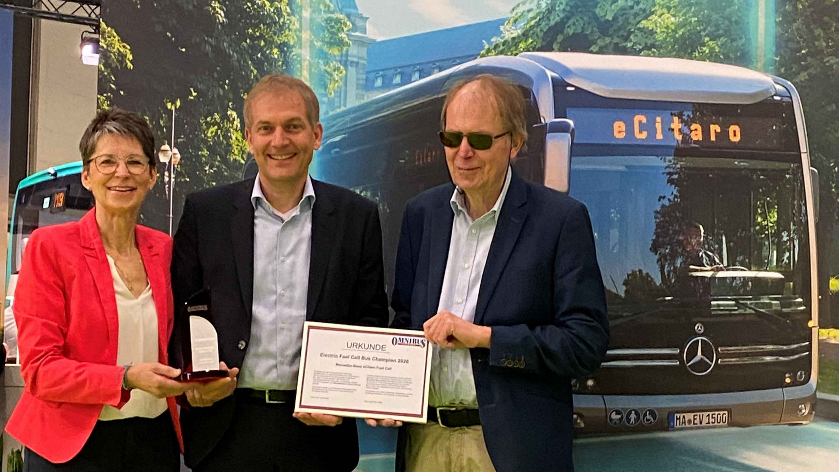 Three people holding a certificate and award in front of a Mercedes-Benz eCitaro in the background