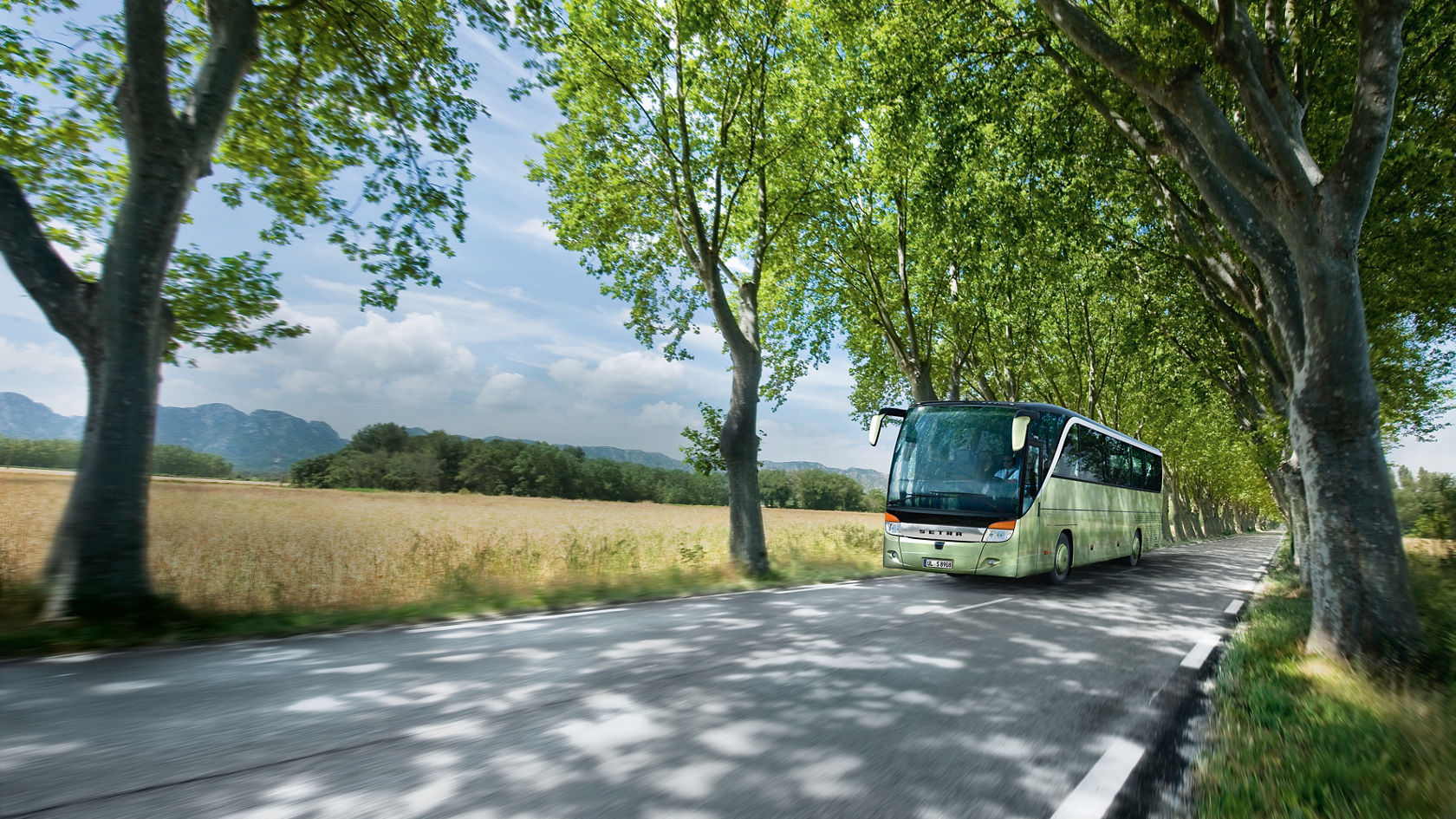 Light green Setra TopClass touring coach driving on a tree-lined country road along fields and hills
