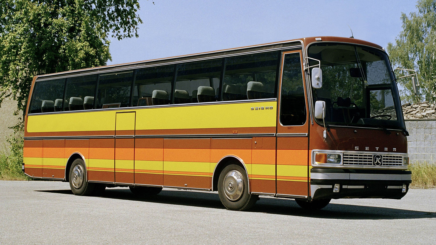 Historic Setra S 215 touring coach in brown and yellow, side view on a road in front of trees and walls