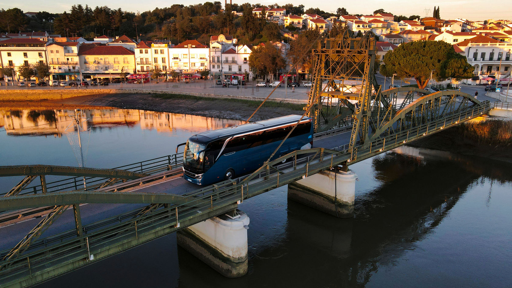 Setra TopClass touring coach drives over a steel bridge over a river in evening light, behind it a shoreline promenade and old town