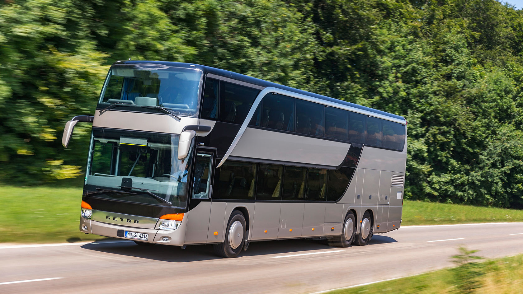 Silver-coloured Setra double-decker bus driving on a country road, side view from the front at an angle against a green forest background