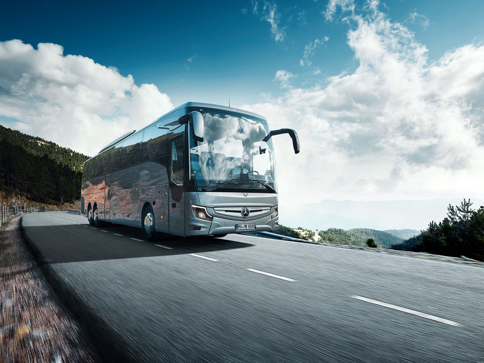 Mercedes-Benz Tourismo Reisebus auf einer Landstraße in den Bergen, mit Waldhang und weitem Himmel mit Wolken