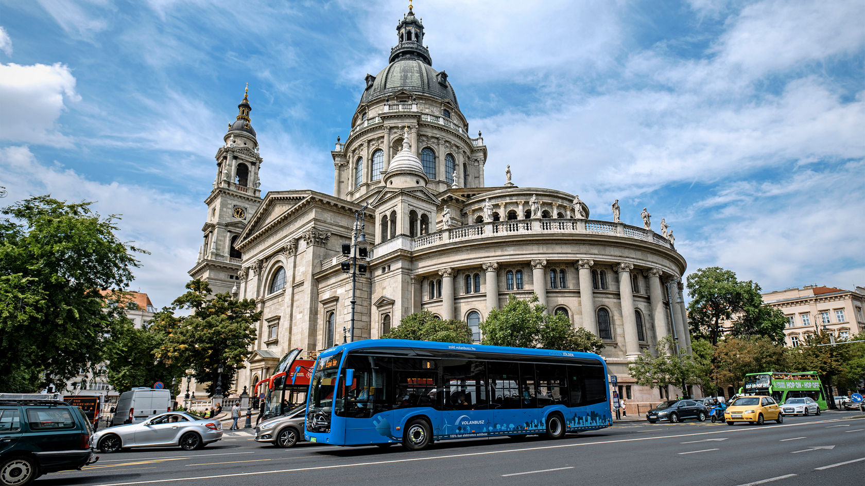 Niebieski autobus miejski Mercedes-Benz eCitaro jedzie w ruchu miejskim wielopasmową drogą przed historycznym kościołem kopułowym 