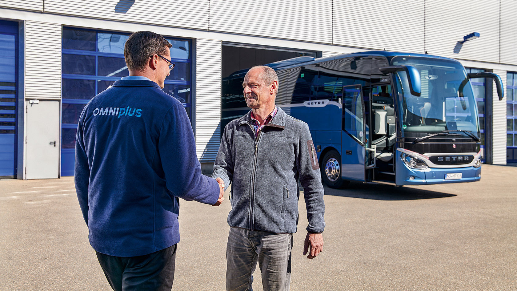 An OMNIplus service employee and another person standing in front of a factory hall shaking hands, a parked Setra ComfortClass bus in the background.