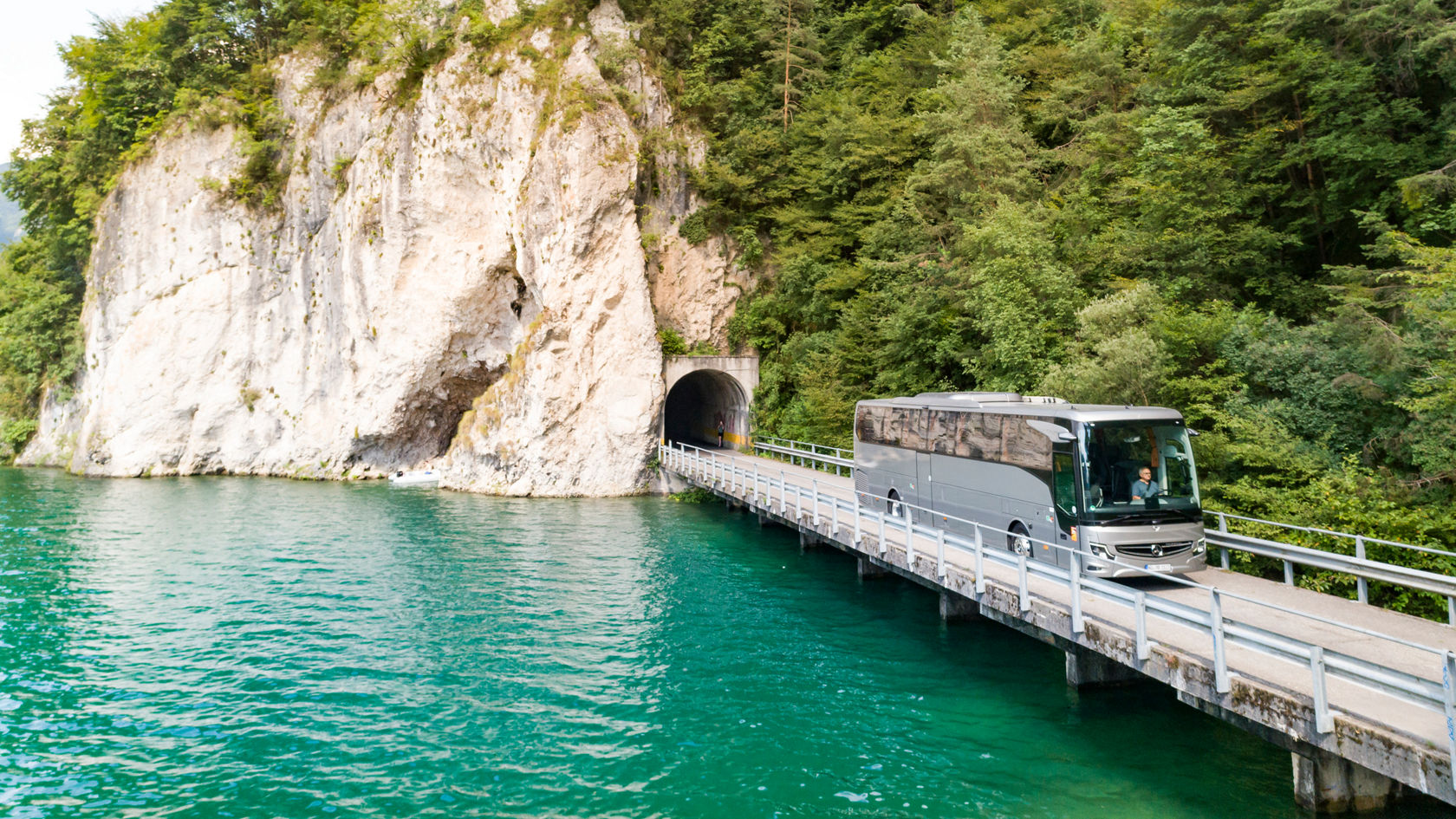 Mercedes‑Benz Tourismo Reisebus fährt auf schmaler Brücke am türkisfarbenen See entlang, vor Tunnel im Fels