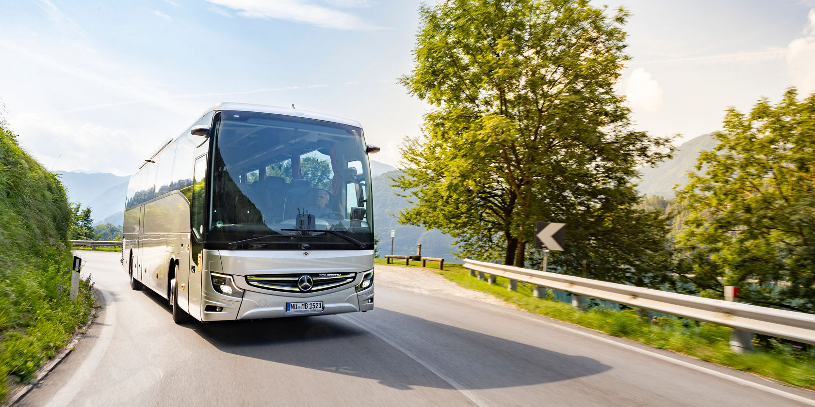 Mercedes‑Benz Tourismo Reisebus fährt auf kurviger Straße in bergiger Landschaft, Leitplanke und Baum am Straßenrand