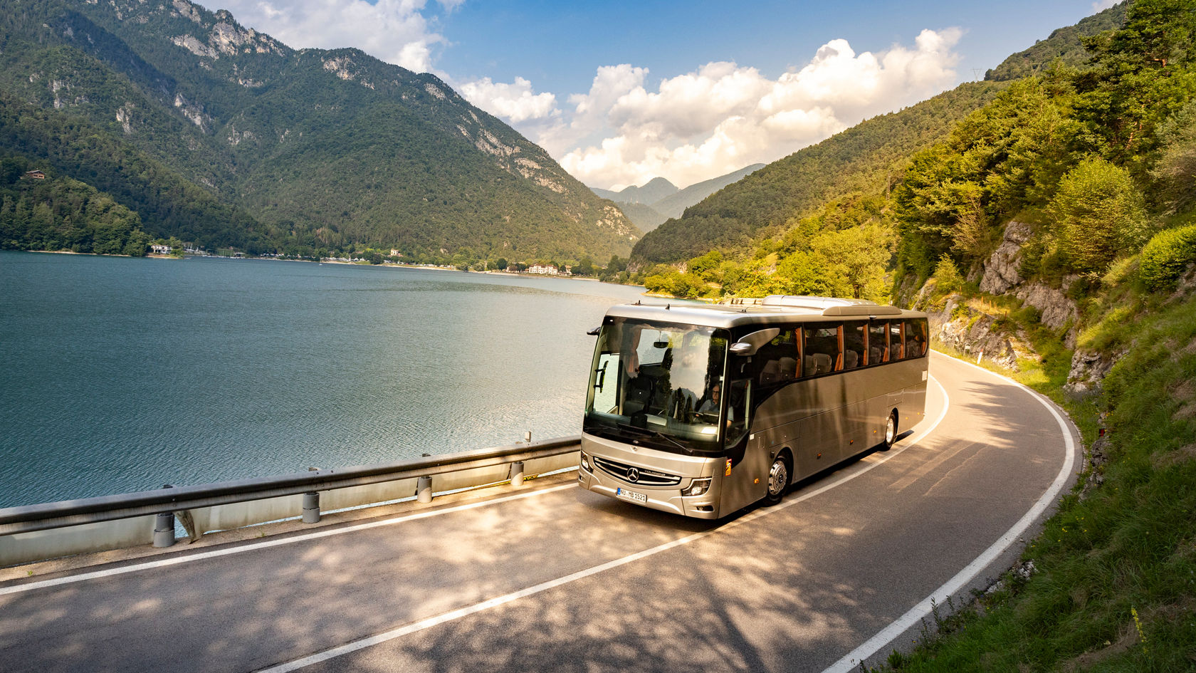 Mercedes‑Benz Tourismo Reisebus fährt auf kurviger Straße am Bergsee entlang, umgeben von grünen Bergen und Wolken