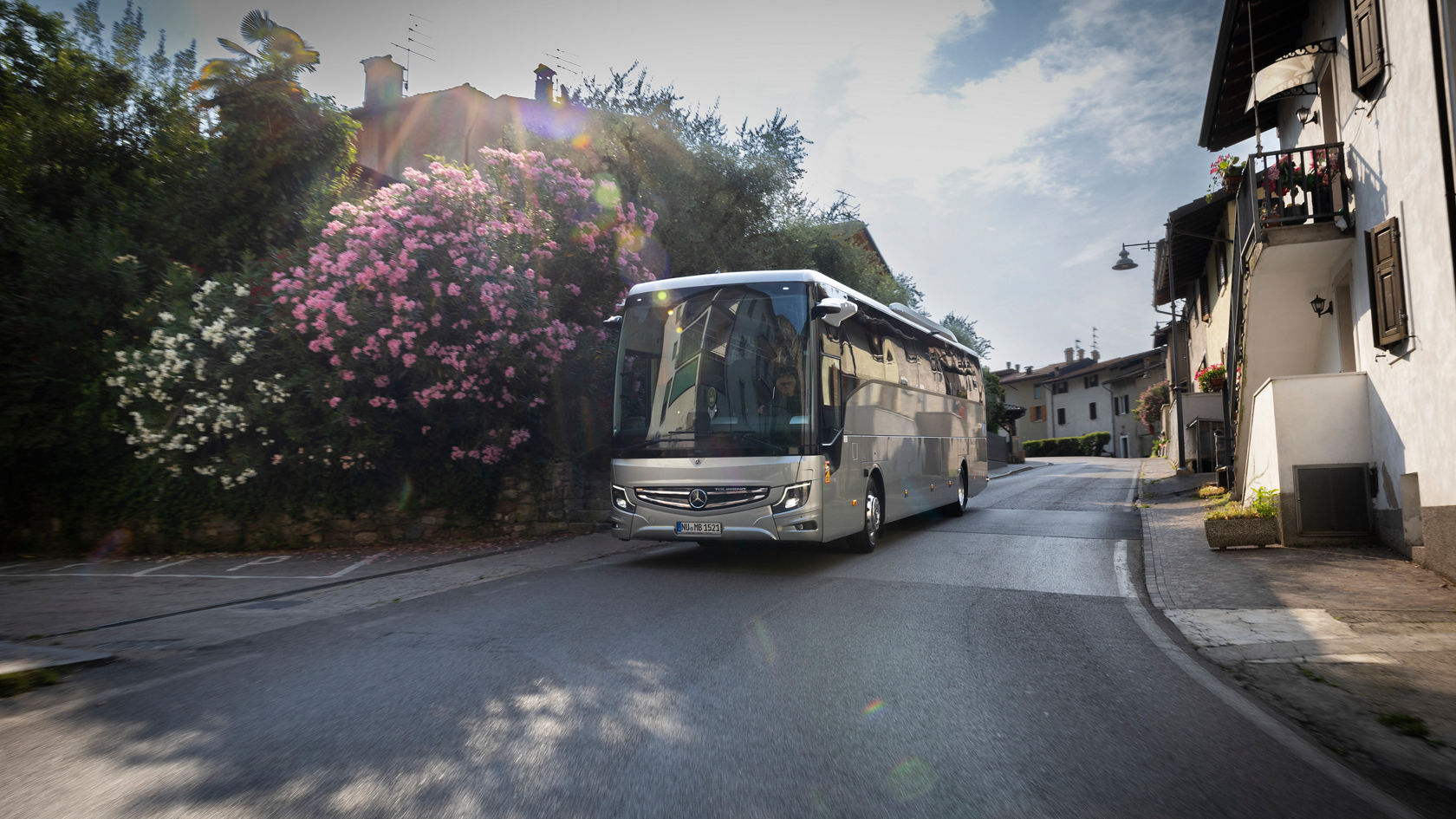 Mercedes‑Benz Tourismo Reisebus fährt durch eine schmale Ortschaftsstraße, vorbei an Häusern und blühenden Sträuchern
