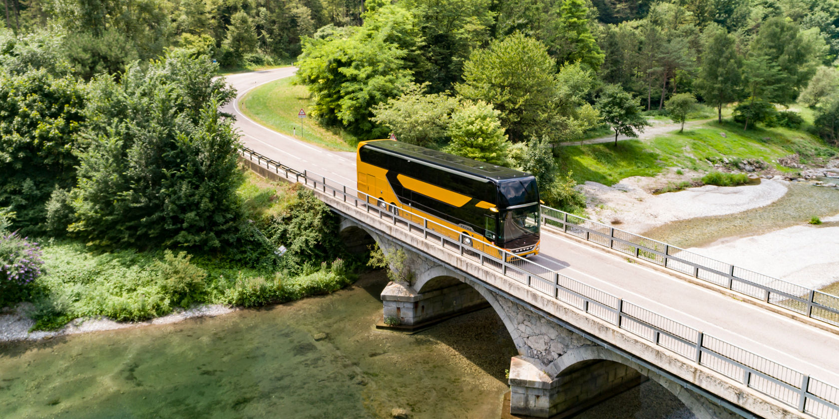 Setra Doppelstock Reisebus überquert eine Brücke über einen klaren Fluss, umgeben von Wald und kurviger Landstraße, Ansicht aus der Vorgelperspektive