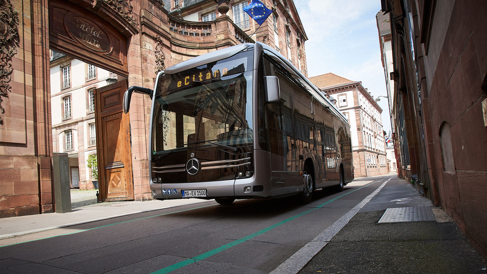 Mercedes-Benz eCitaro Stadtbus fährt durch eine historische Altstadtstraße, im Hintergrund ein Sandstein-Torbogen