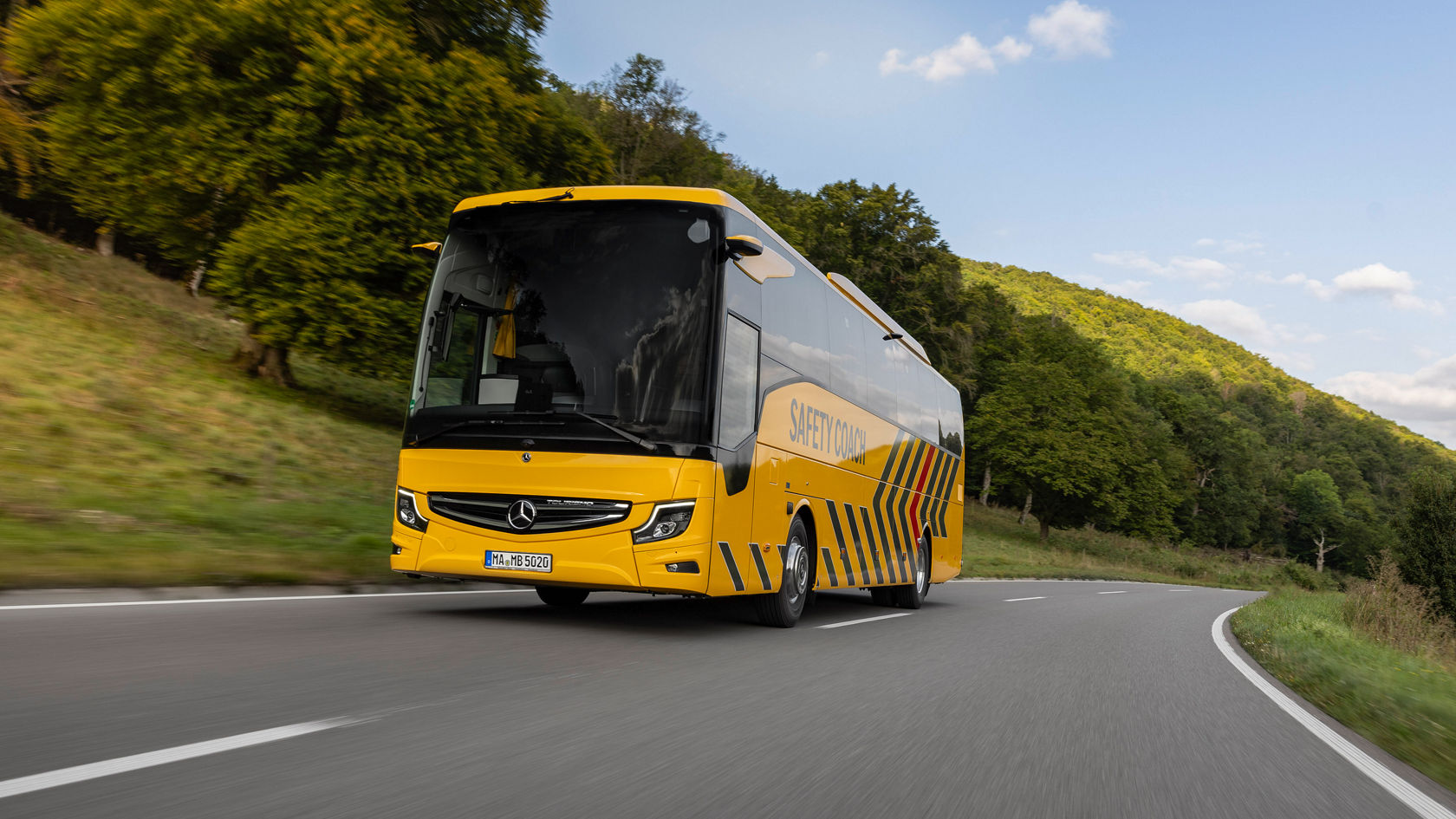 Yellow Mercedes‑Benz Tourismo “Safety Coach” touring coach driving on a winding country road through a forested hilly landscape