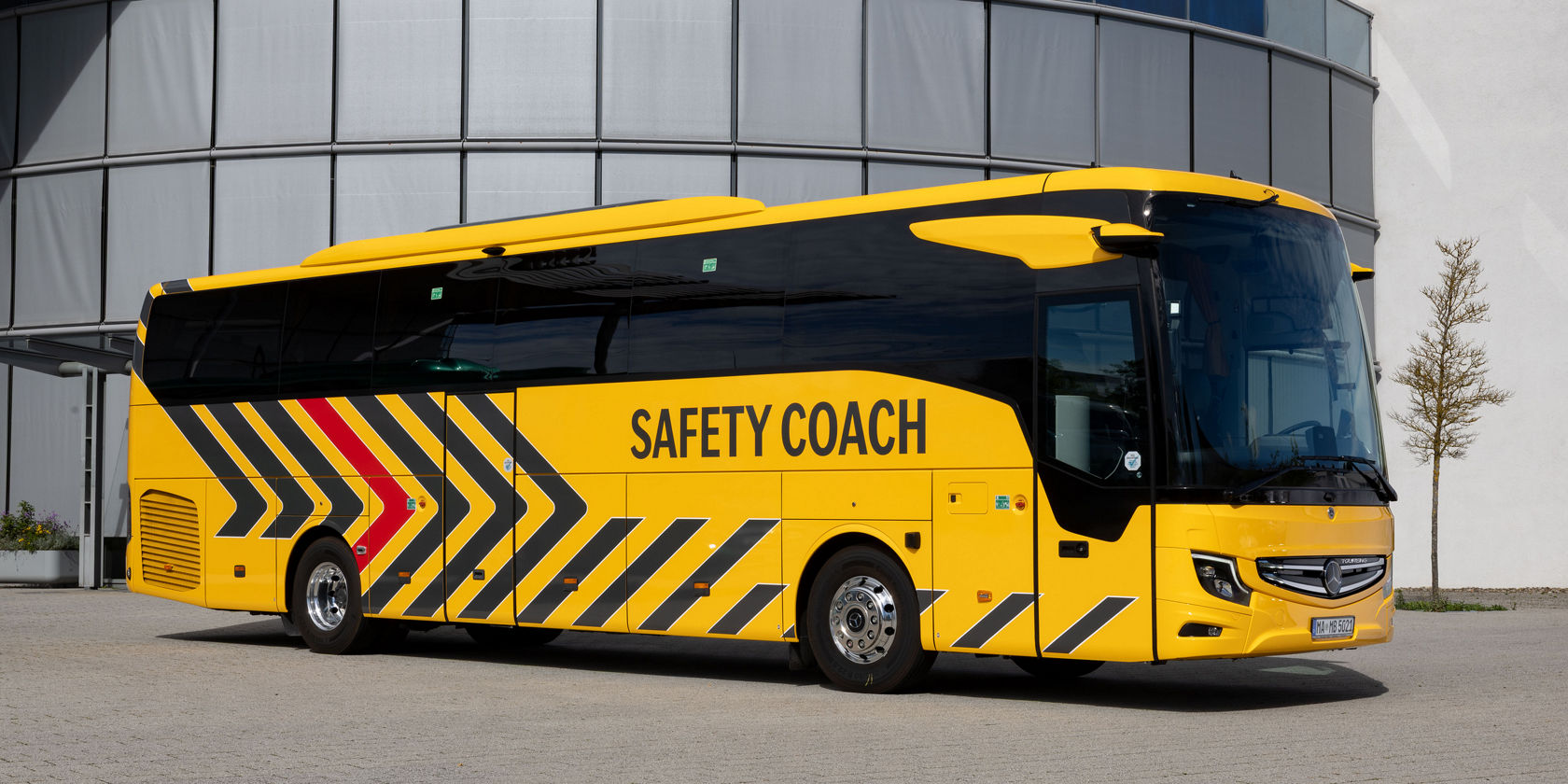 Gelber Mercedes‑Benz Tourismo Safety Coach mit Markierungen steht vor modernem Gebäude, Seitenansicht