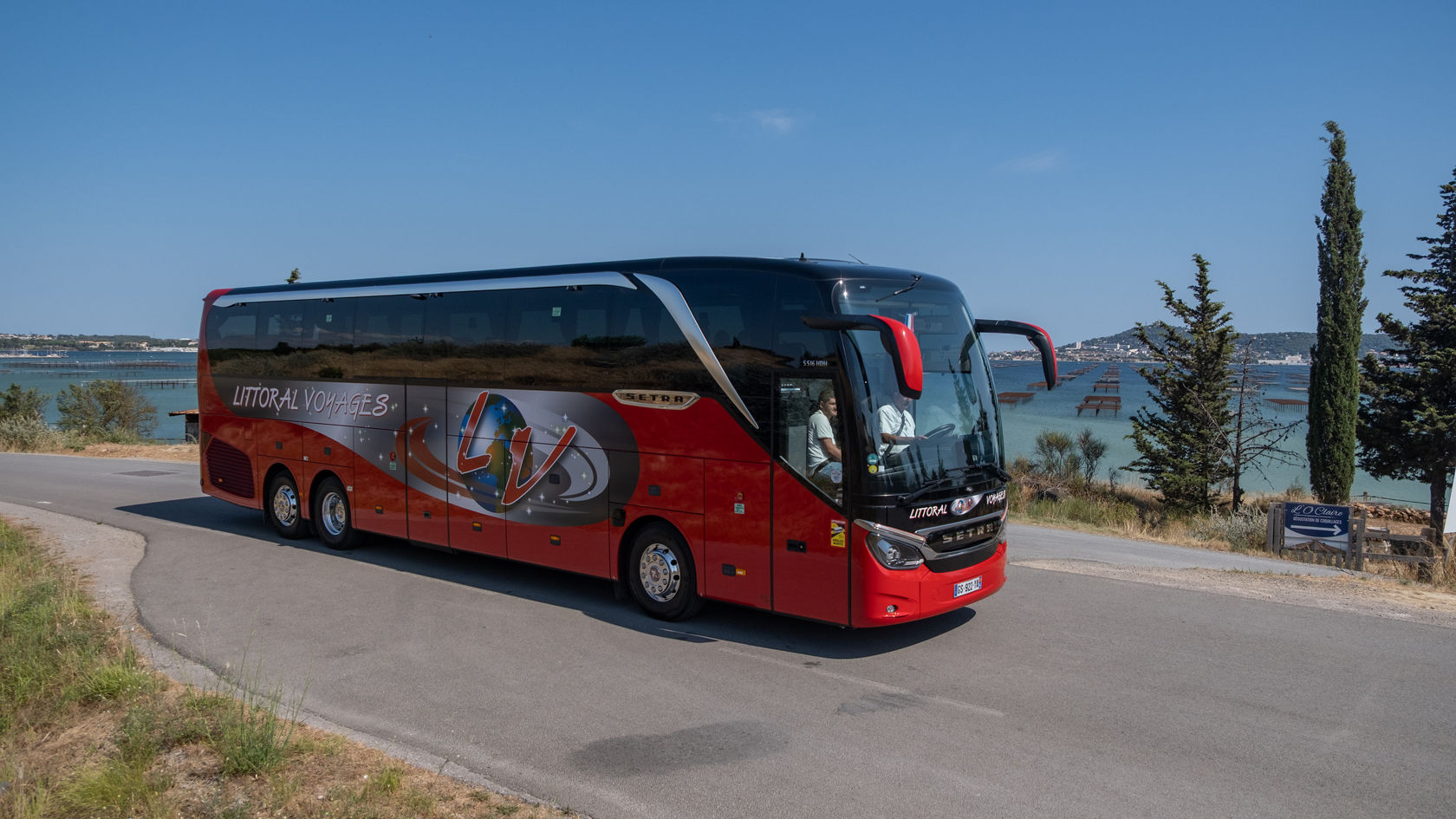 Red Setra TopClass touring coach driving on a coastal road along a lagoon, with water and shore landscape in the background