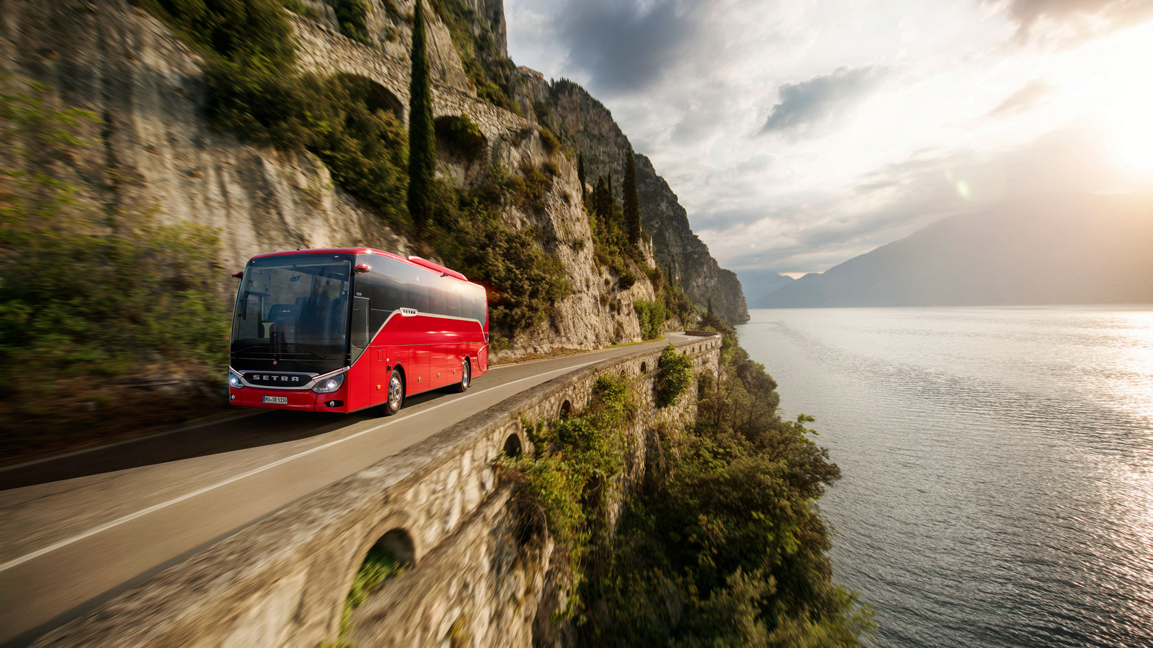 Red Setra ComfortClass touring coach driving on a winding coastal road along steep rocks, with water on the right in the backlight
