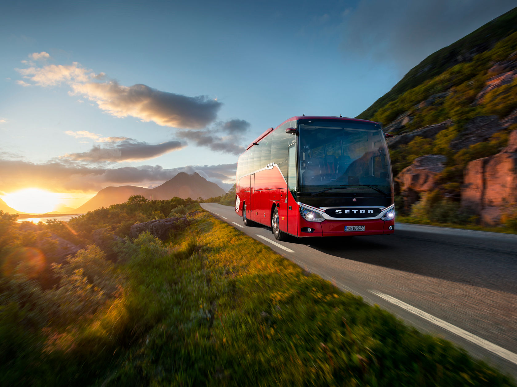 Autocar Setra ComfortClass rojo circulando por una carretera costera al atardecer, con montañas y mar al fondo