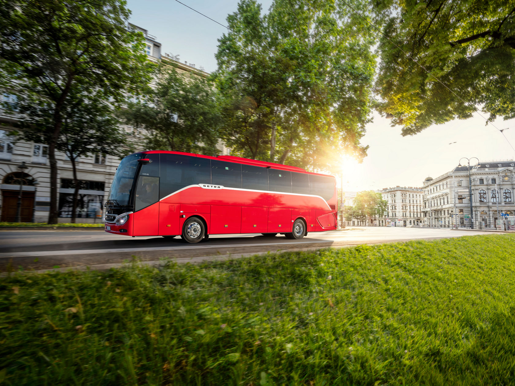Autocar Setra ComfortClass rojo circulando por una avenida urbana con el sol bajo, con árboles y edificios históricos al fondo.