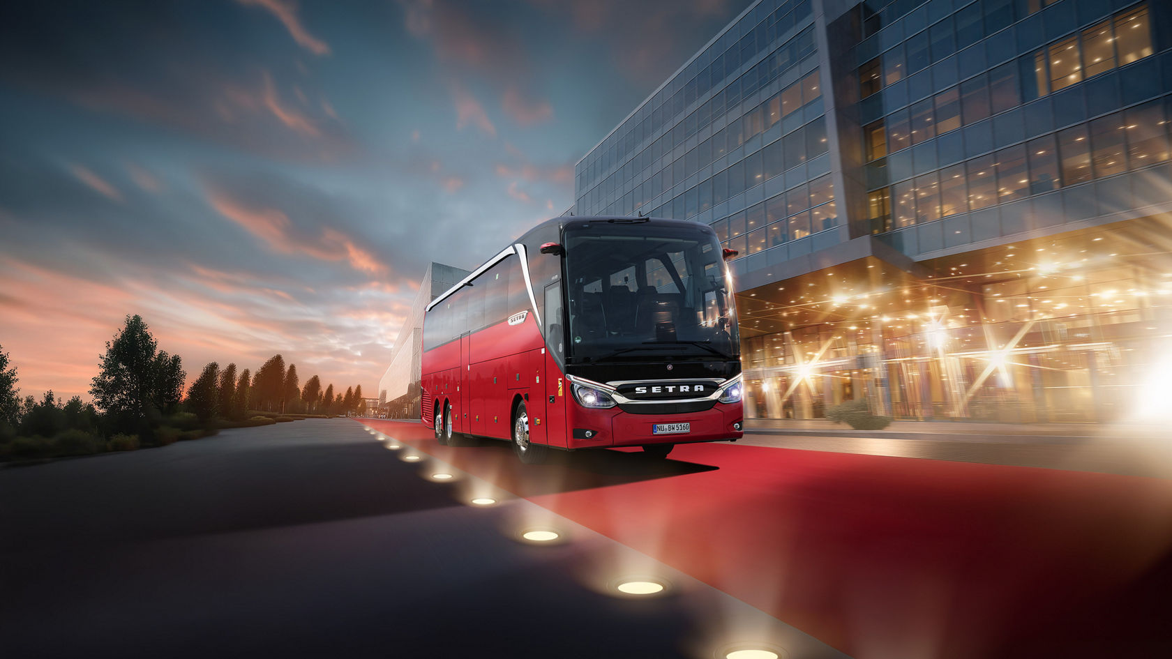 Red Setra TopClass touring coach in front of modern glass facade at dusk, on red carpet with floor lights in front of the entrance