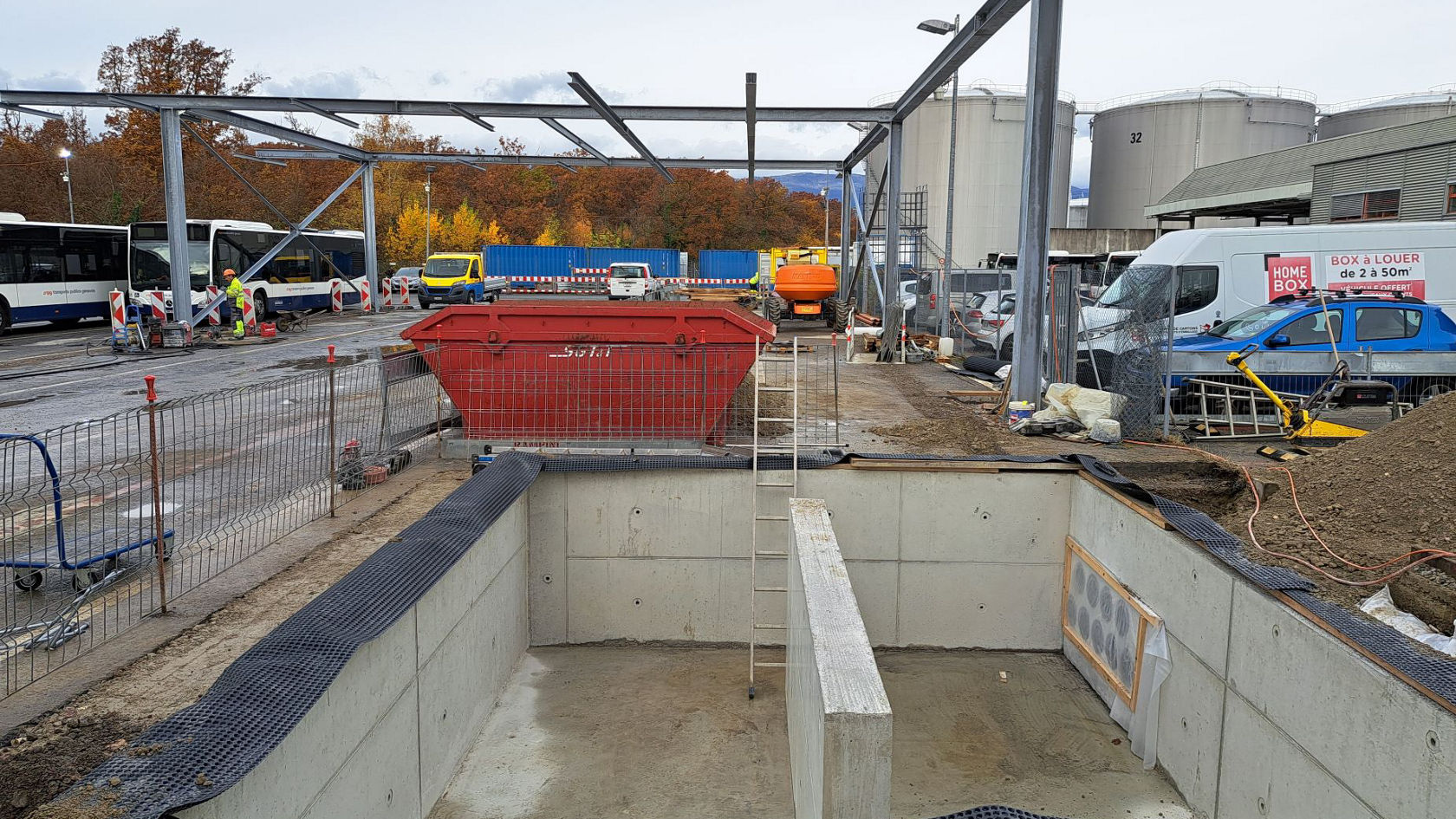 Baustelle für neue Ladeinfrastruktur auf einem Betriebshof: offener Betonbaugraben, Stahlkonstruktion im Aufbau und Busflotte im Hintergrund für zukünftige Elektrobus‑Ladestationen mit Mega-Charger Geräten