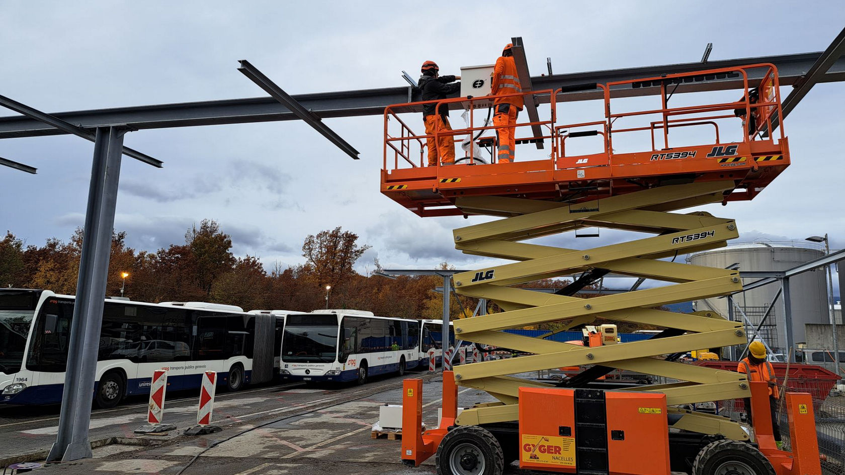 Arbeiter montieren auf einer Hebebühne Stahlträger für die neue Ladeinfrastruktur eines Busbetriebshofs; im Hintergrund stehen mehrere Mercedes‑Benz Stadtbusse.