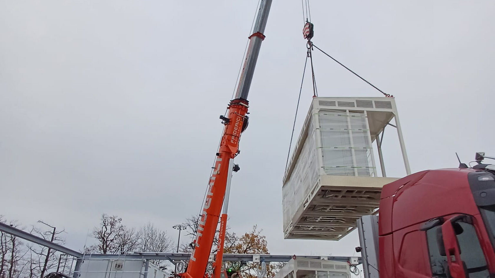 Ein Kran hebt ein großes Technikmodul für die Ladeinfrastruktur auf einem Busbetriebshof an; Schwerlastkran und Lkw arbeiten gemeinsam beim Aufbau moderner Elektrobus‑Ladestationen.