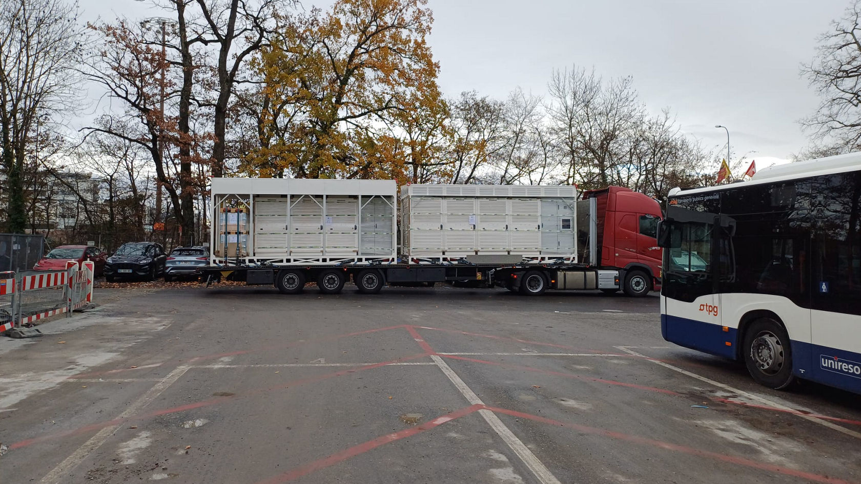 Ein Lkw liefert große Technikmodule für die Ladeinfrastruktur auf einen Busbetriebshof; im Vordergrund steht ein Mercedes‑Benz Stadtbus der TPG, im Hintergrund herbstliche Bäume.