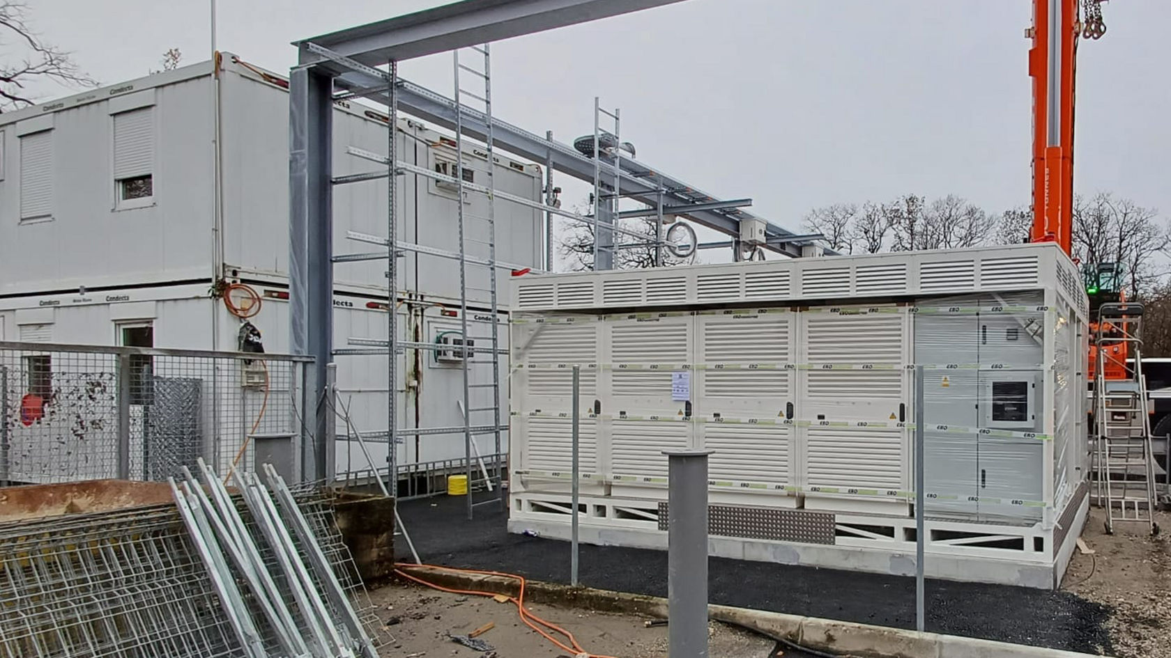 Baustelle für Ladeinfrastruktur auf einem Busbetriebshof: weißes Technikmodul, umgeben von Stahlkonstruktion und Baugeräten für zukünftige Elektrobus‑Ladestationen.