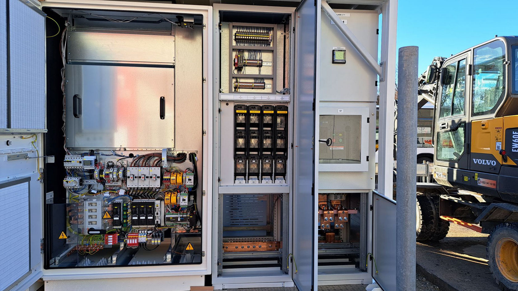 Geöffneter Schaltschrank einer Elektrobus‑Ladeinfrastruktur mit sichtbaren Leistungsschaltern, Steuertechnik und Verkabelung auf einer Baustelle; daneben steht ein Bagger.