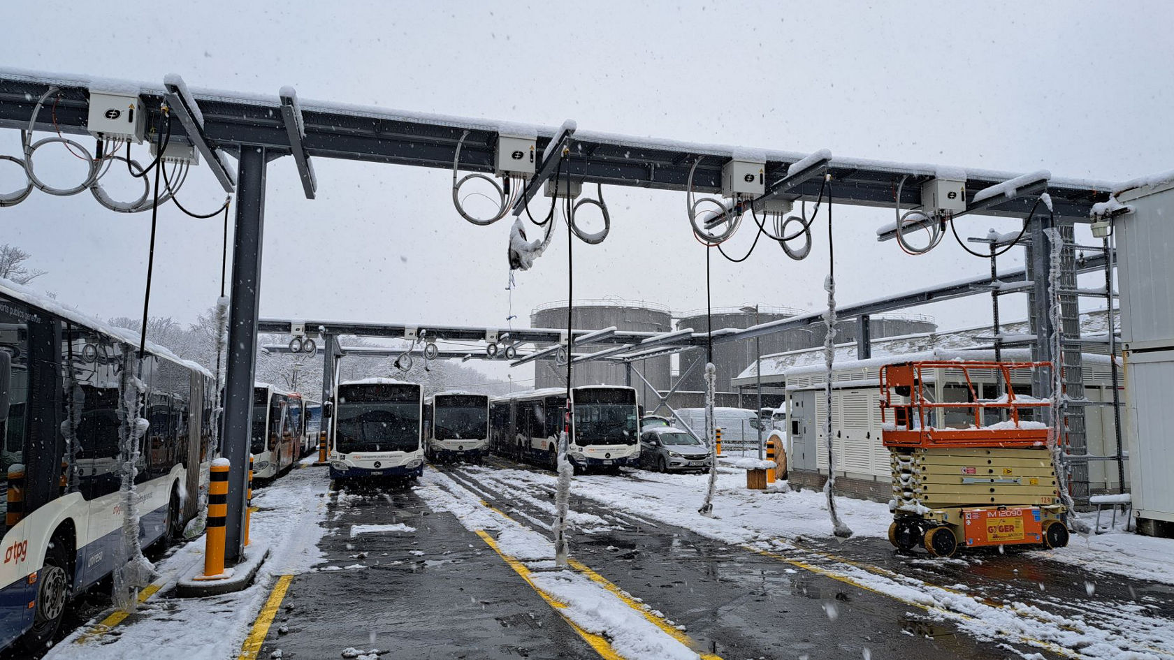 Mehrere Mercedes‑Benz Stadtbusse stehen im verschneiten Depot unter einer technischen Lade‑ und Serviceinfrastruktur; winterliche Szenerie mit Neuschnee auf Fahrzeugen, Anlagen und Überdachung.