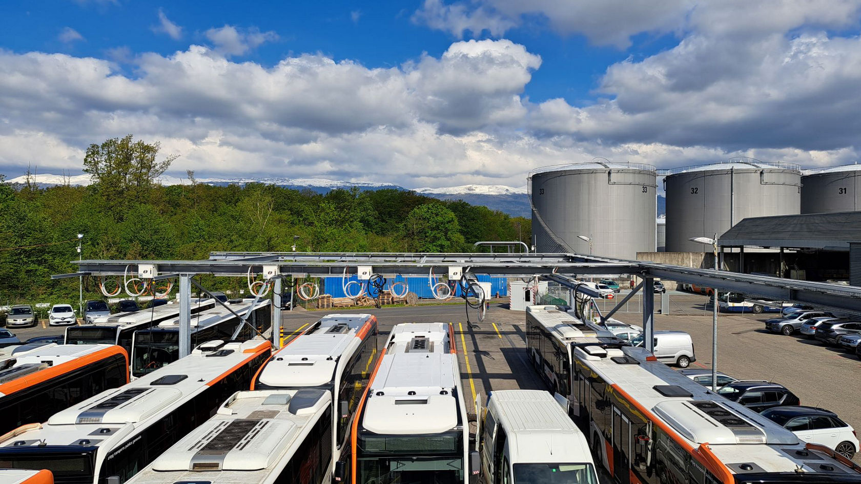 Mehrere Mercedes‑Benz Elektro‑ und Stadtbusse stehen auf einem Betriebshof unter einer Ladeinfrastruktur; im Hintergrund Industrieanlagen und eine bewaldete Landschaft mit Bergen.