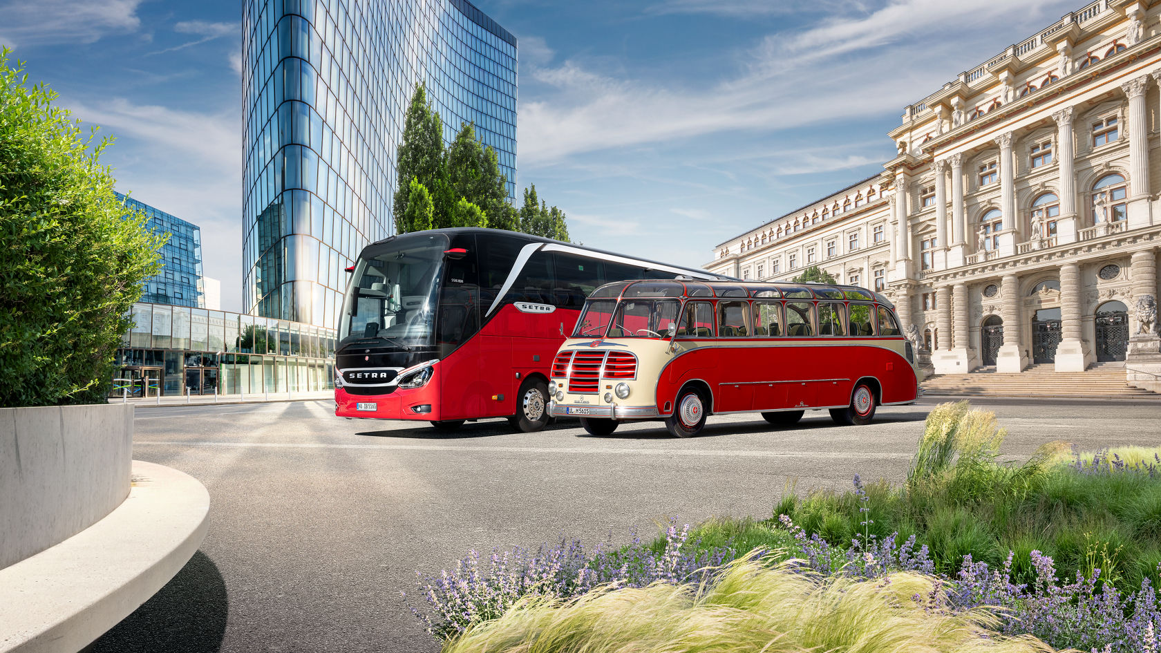 A modern red Setra TopClass S516 touring coach stands next to a historic Setra classic bus, with a modern and classic building ensemble in the background.