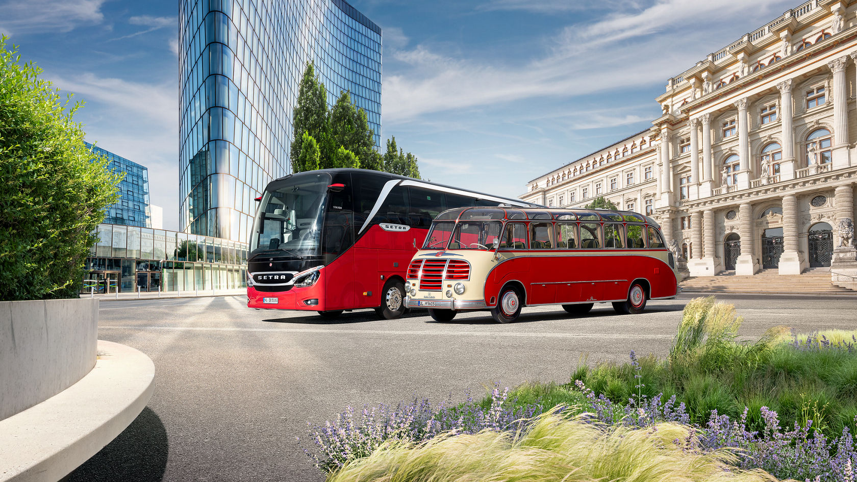 A modern red Setra TopClass S516 touring coach stands next to a historic Setra classic bus, with a modern and classic building ensemble in the background.