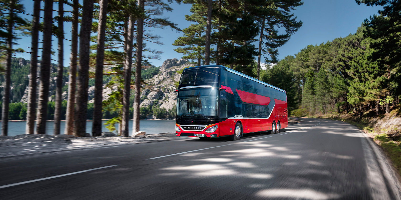 Roter Setra S531 Doppelstock-Reisebus fährt auf Landstraße am See entlang, umgeben von Kiefern und Bergen im Hintergrund