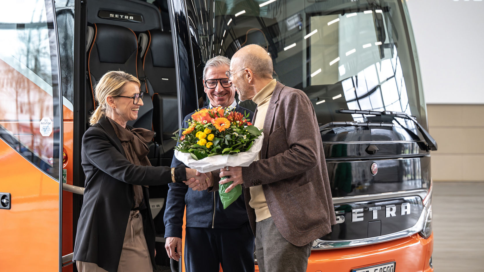Tre persone davanti a un autobus da turismo Setra nel Centro Clienti, una persona consegna un mazzo di fiori in presenza dell'altra.