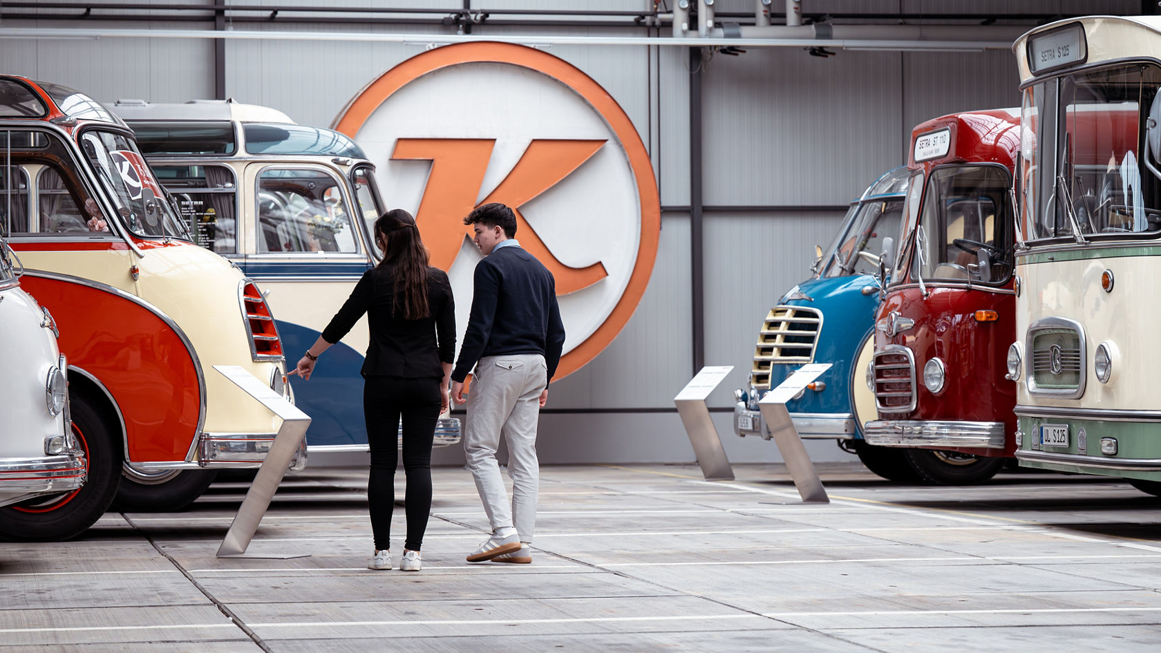 Twee bezoekers in de Kässbohrer-hal tussen historische Setra bussen, lezen informatieborden in de tentoonstelling