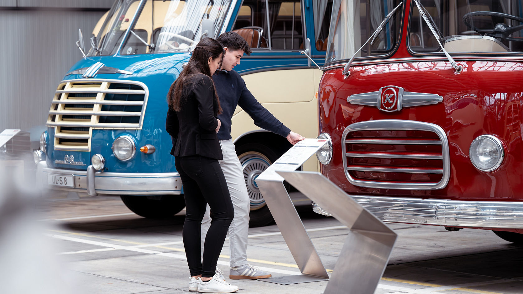 Twee bezoekers lezen in de Kässbohrer-hal een informatiebord voor historische Setra touringcars in blauw en rood
