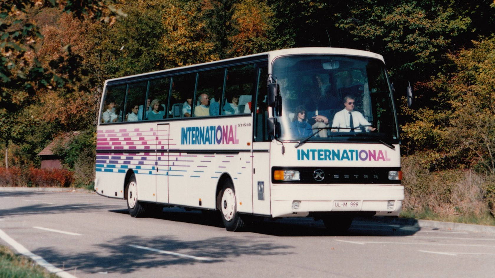 Setra touring coach with “International” lettering driving on country road, side view in front of autumnal forest
