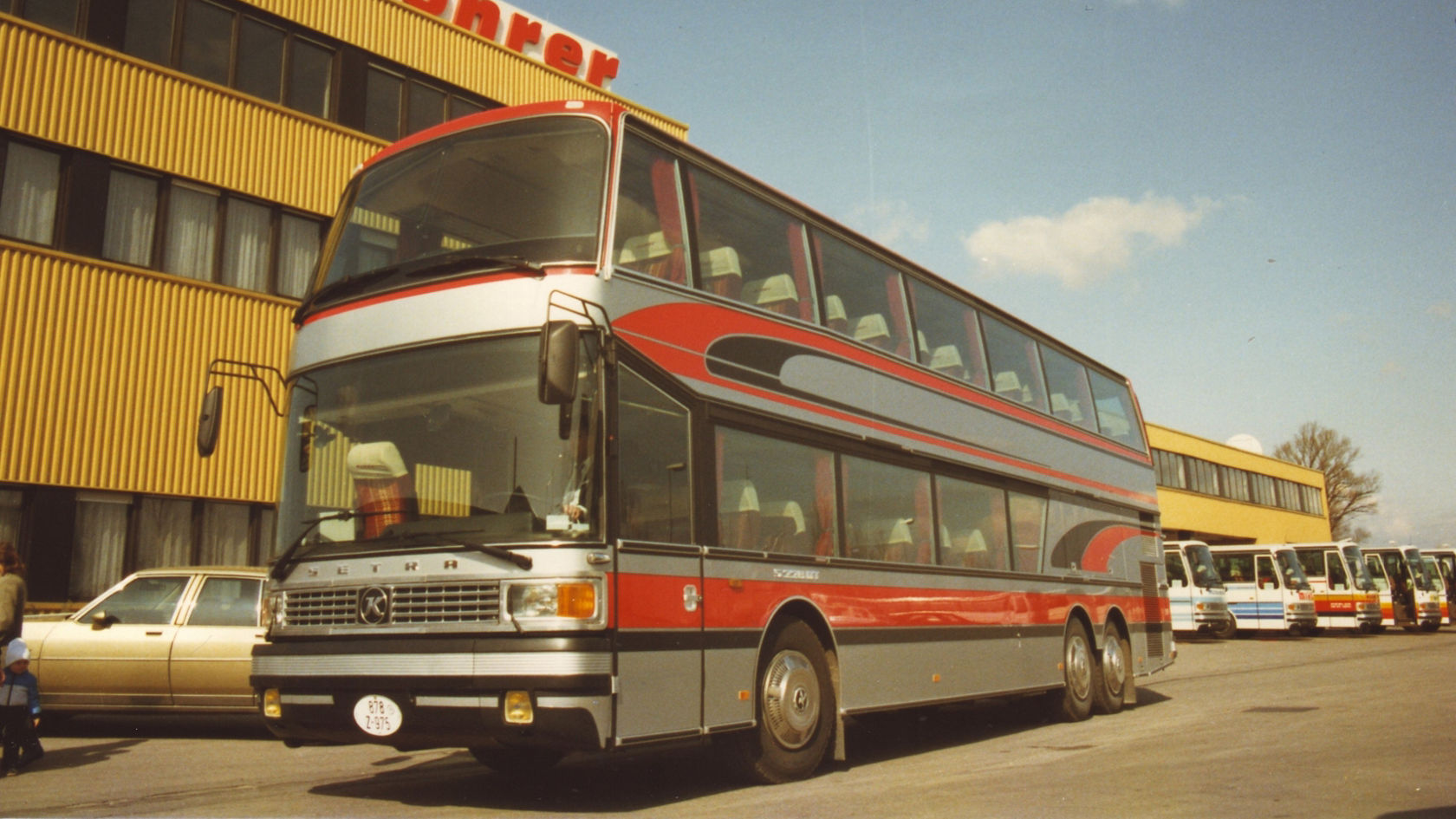 Setra double-decker coach in red and silver in front of a company building with Kässbohrer lettering on the roof, other buses in the yard in the background