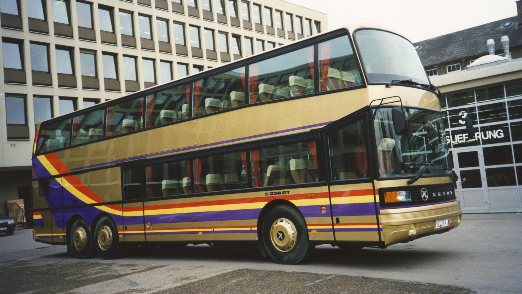 Setra double-decker coach in gold with colourful stripes, parked at an angle from the front in front of an office building