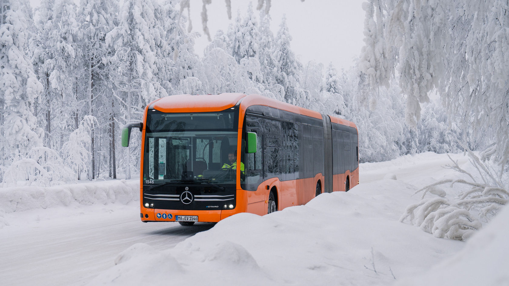Orange Mercedes-Benz eCitaro articulated bus driving on a snow-covered road through a wintery forest