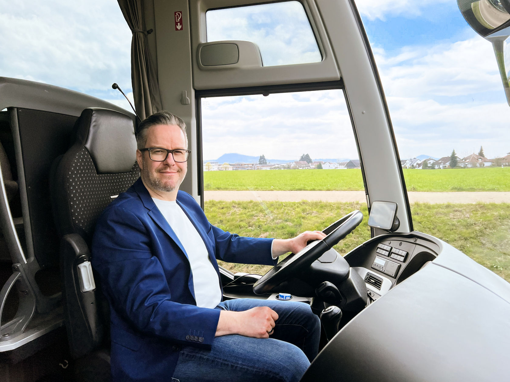 El conductor está sentado en el puesto de conducción de un autocar Setra ComfortClass, mirada lateral al caballero en la entrada, en segundo plano una gran ventanilla lateral con campos verdes 