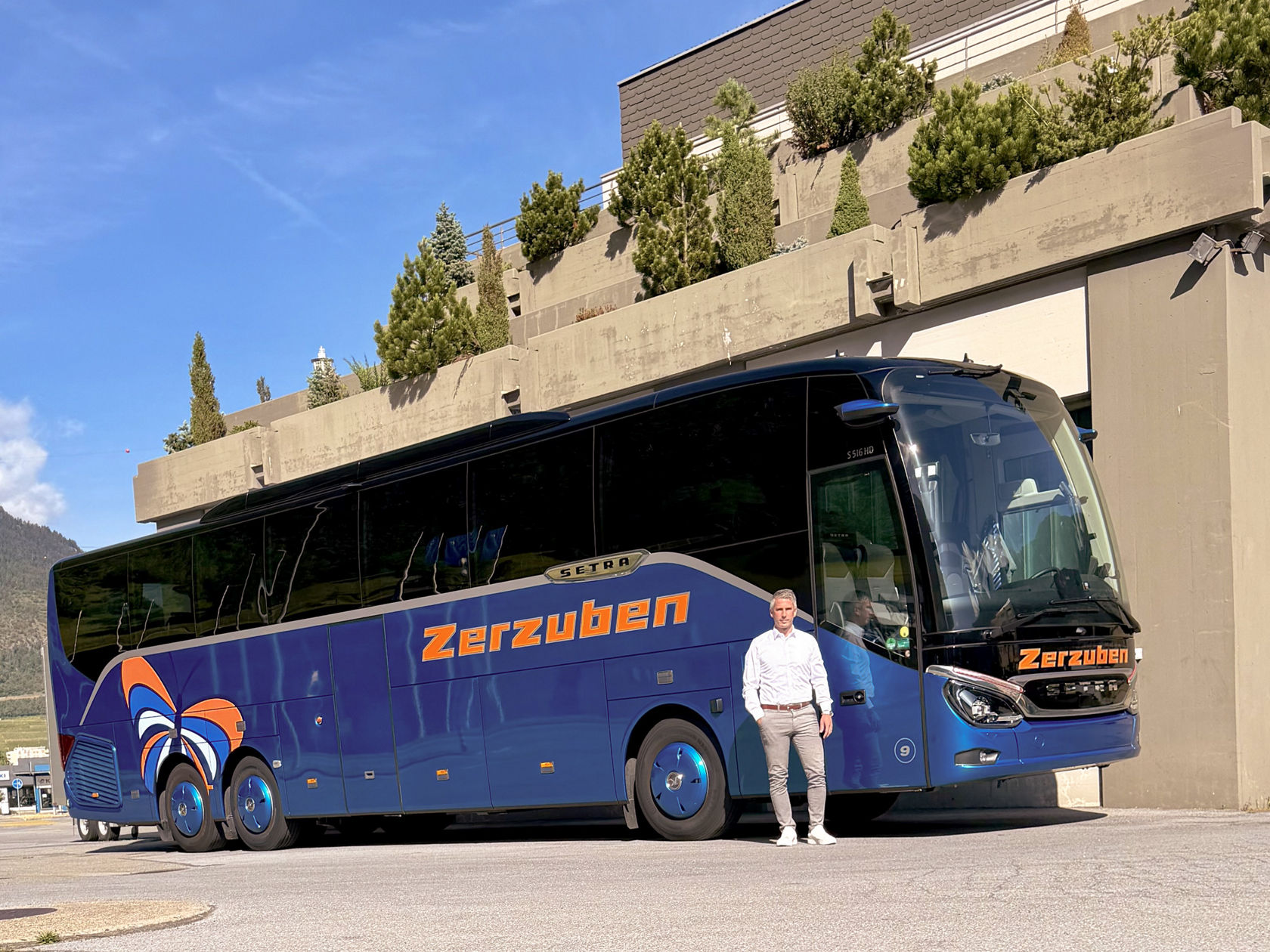 Autocar Setra azul delante de un edificio de varias plantas con balcones plantados bajo el sol, delante de él una persona en primer plano 