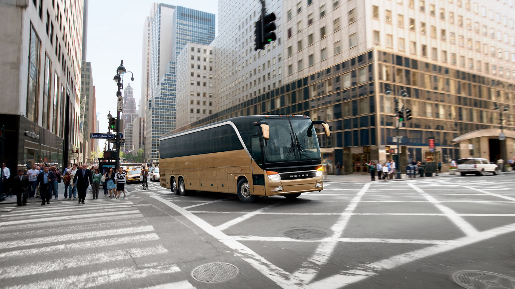 Gold-coloured Setra TopClass touring coach drives through a bustling American city centre intersection with high-rise buildings and pedestrians