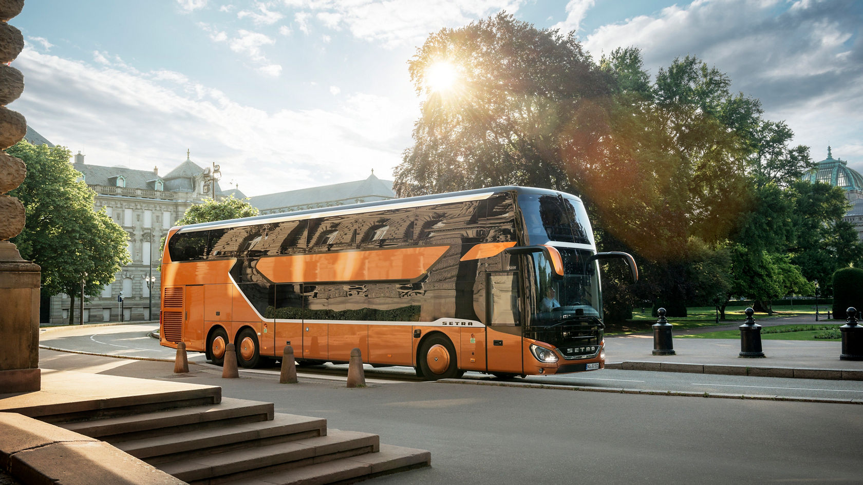 Orange Setra double-decker bus standing on the side of the road in front of historic buildings, sun shining through trees