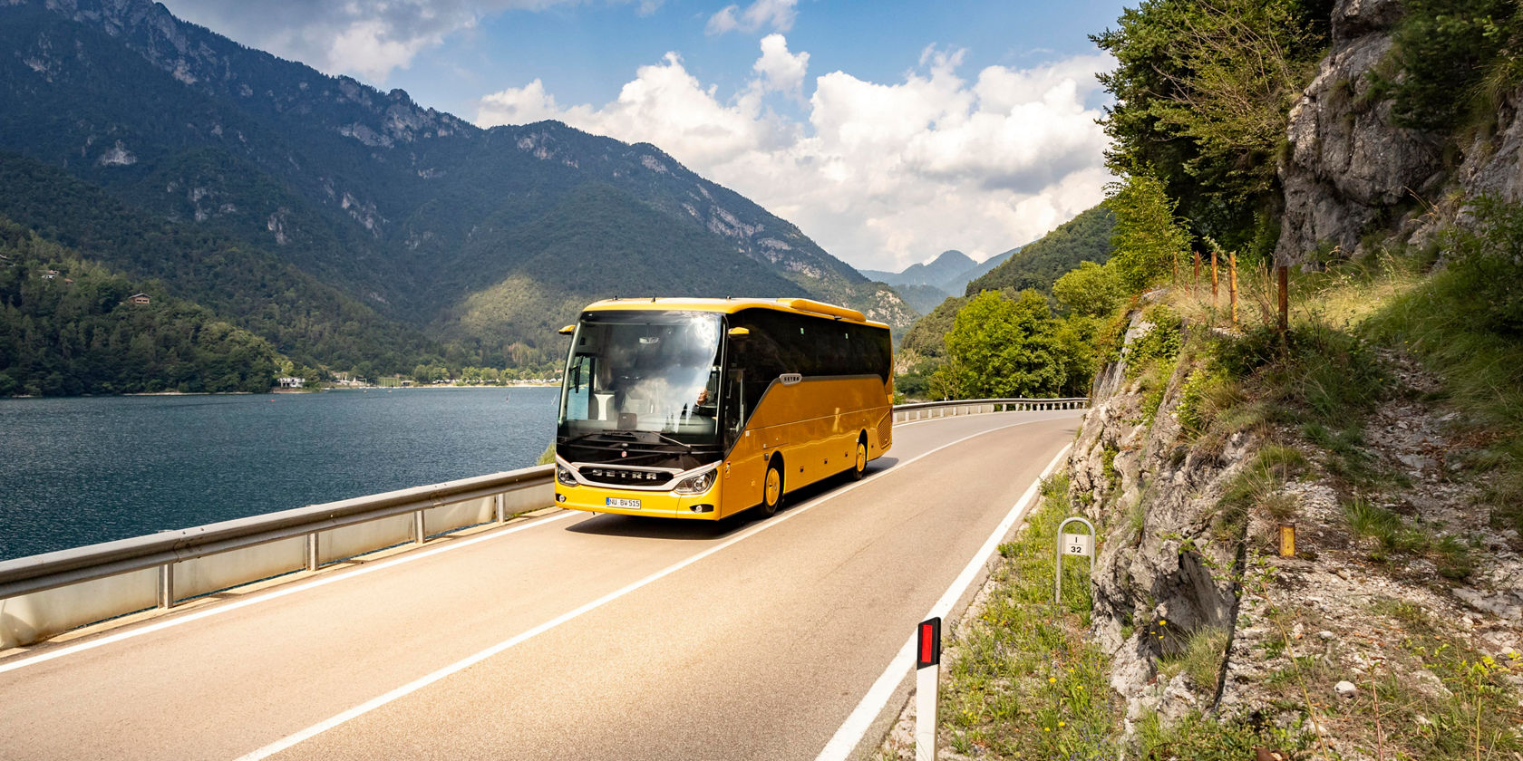 Autocarro de turismo Setra ComfortClass amarelo a circular numa estrada costeira junto ao lago, com montanhas e parede rochosa em segundo plano