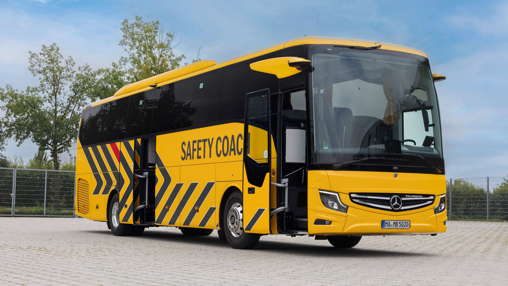 Gelber Mercedes‑Benz Tourismo Reisebus „Safety Coach“ mit geöffneten Türen, Seitenansicht auf gepflastertem Parkplatz