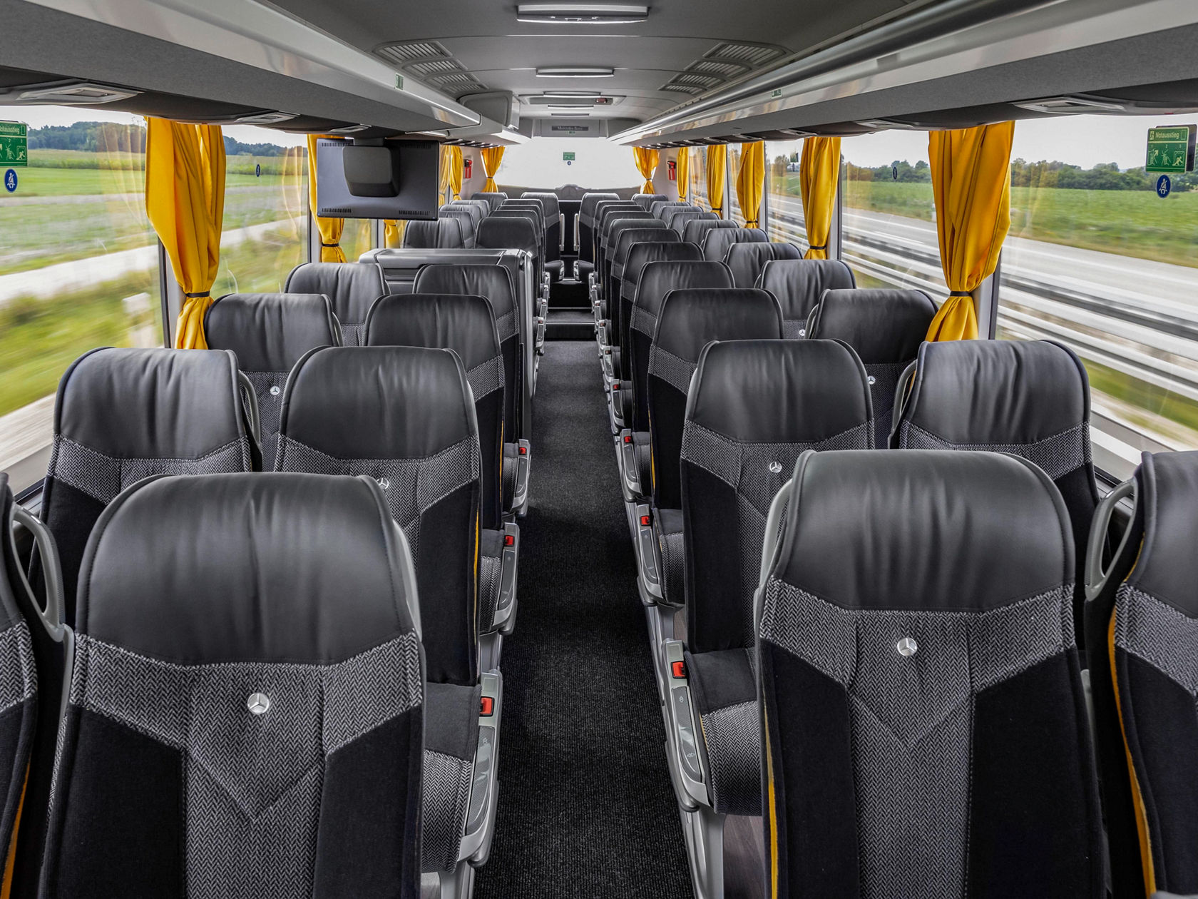 Innenraum eines Mercedes-Benz Tourismo Reisebusses mit Reihen schwarzer Sitze und gelben Vorhängen, Blick nach vorn durch den Mittelgang