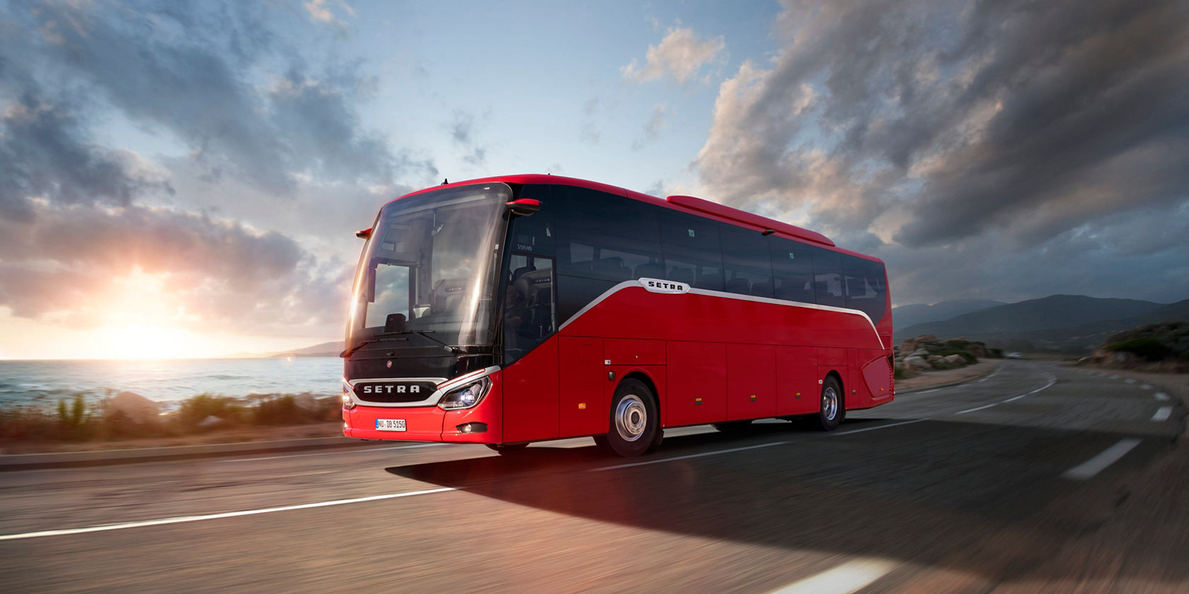 Autocarro de turismo Setra ComfortClass vermelho a circular numa estrada costeira ao pôr do sol, mar e céu nublado em segundo plano