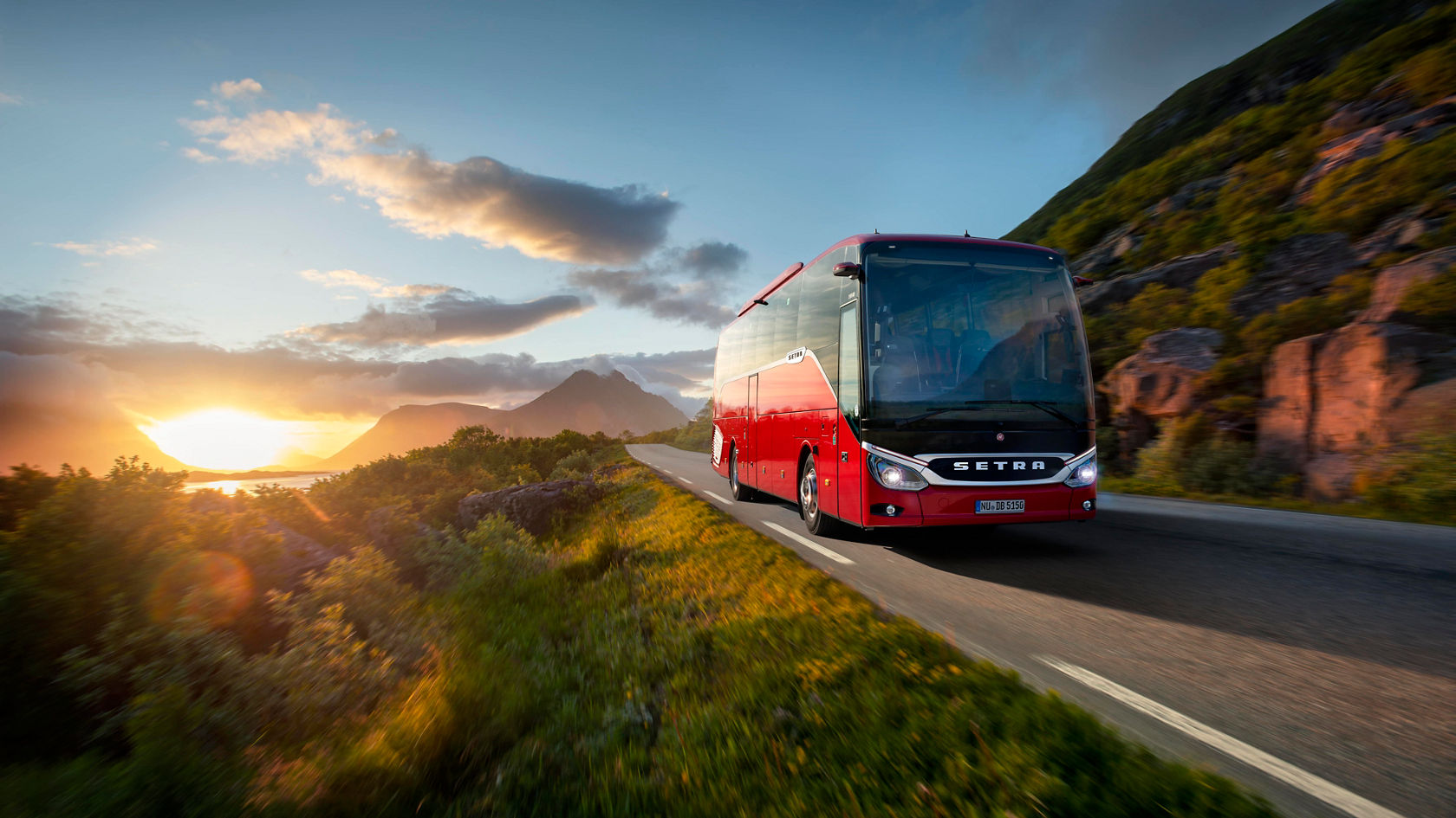 Autocarro rodoviário Setra ComfortClass vermelho a circular ao pôr do sol numa estrada costeira, com montanhas e mar em segundo plano