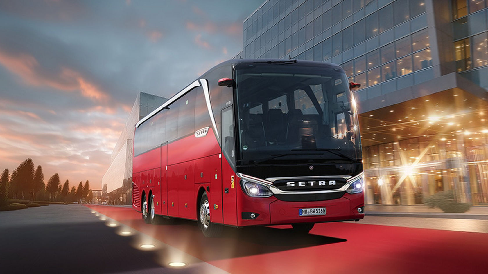 L'autobus da turismo rosso Setra TopClass viaggia al crepuscolo su un tappeto rosso lungo un moderno edificio in vetro, vista frontale con faro sinistro e destro