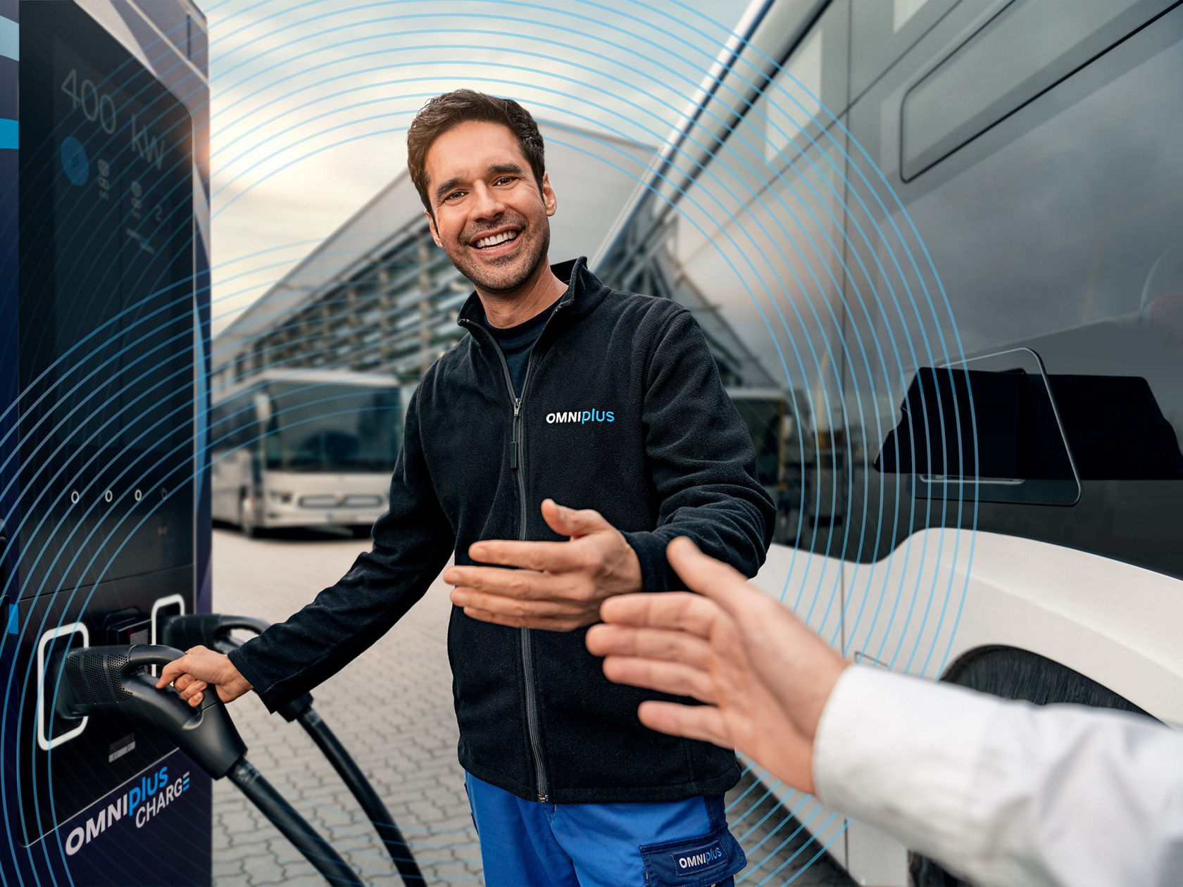 Pracownik OMNIplus przy terminalu ładowania OMNIplus CHARGE, wkłada przewód do ładowania do autobusu elektrycznego i wita klienta dłonią